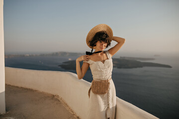 Wall Mural - Attractive young brunette woman in beige dress and straw hat poses in beautiful place with sea view. Portrait of curly lady in elegant outfit outside.