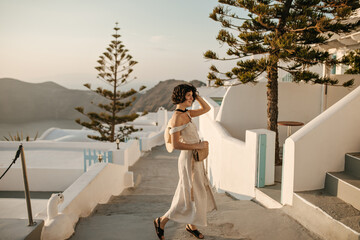 Curly brunette woman in midi beige dress moves and walks in beautiful place with white buildings. Charming lady with straw bag poses outside.