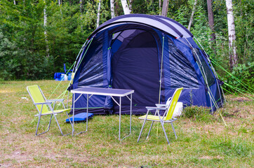 Installed blue tourist tent, camping table and chairs in forest at campsite.