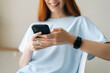 © dikushin - Close-up of smiling young woman holding smart phone looking on display screen, using smartphone texting online message sitting in light room. Pretty redhead Caucasian lady having leisure activity.