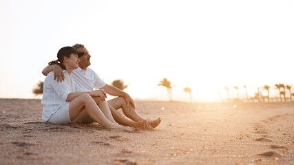 Wall Mural - Happy mature senior couple sitting on the beach at sunset time. Aging together and retirement age lifestyle concept                              