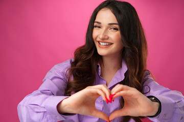 Wall Mural - Cheerful smiling young woman showing heart sign against pink background