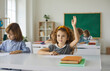 © Studio Romantic - Elementary school student raises her hand, ready to answer the teacher's questions in class. Smart little curly girl is sitting at a desk next to her classmate in the classroom. Concept of education.