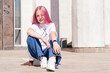 © Tatyana - A pink-haired hipster teenage girl in a white T-shirt and jeans is sitting on a skateboard on a city street.Generation Z style,active lifestyle,summer concept.Copy space for text.