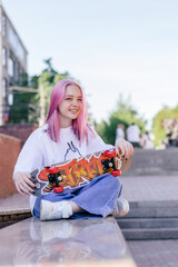 Positive pink-haired hipster teenage girl in a white T-shirt holding a bright skateboard on a city street on a summer day.Generation Z style,active lifestyle.Copy space.