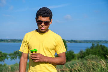 Wall Mural - Smiling African American man in sunglasses enjoying the taste of coffee outdoors in summer by the river, looking and standing in front of the camera. Copy space