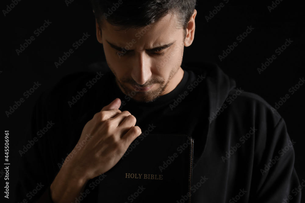 Religious young man with Bible praying on dark background - obrazy, fototapety, plakaty 