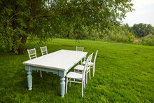 White Table With Chairs Under Tree On Lawn. White Table With Chairs Located On Green Grass Under Lush Tree On Summer Day In Field