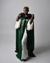 Wall Mural - Full length portrait of pretty African woman wearing long green medieval fantasy gown and velvet cloak, sitting on a light grey studio background.