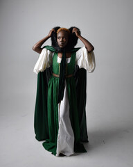 Wall Mural - Full length portrait of pretty African woman wearing long green medieval fantasy gown and velvet cloak, sitting on a light grey studio background.