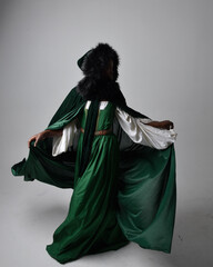 Full length portrait of pretty African woman wearing long green medieval fantasy gown and velvet cloak, sitting on a light grey studio background.