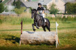 close portrait of handsome rider man jumping over obstacle on black stallion horse during eventing cross country competition in summer