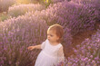 Beautiful babygirl in a white dress walking with a bouquet in a lavender field. Sunset view. Summer scenic view. Portrait of blonde babygirl.