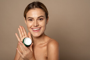 Wall Mural - Portrait of a pretty young smiling woman with clean fresh skin in the studio on a beige background. Cute woman holding a jar of moisturizer in the studio. Skin care and hydration concept