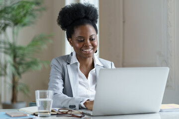 African businesswoman look on laptop screen with happy smile. Young ethnic office worker or boss female reading email on computer with successful good news, communicate with client online at workplace