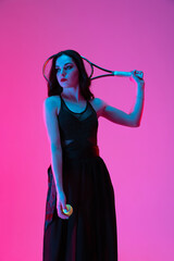 Wall Mural - High-fashion styled young woman in black evening gown, dress playing tennis isolated over pink neon background. Concept of fashion and sport