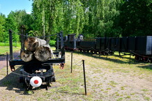 A Close Up On A Railroad Compartment Made Out Of Metal, With Connections Visible And With Some Logs And Planks Loaded Onto It Seen Next To An Old Black Locomotive Seen In The Middle Of A Public Park