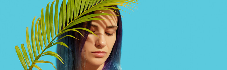 Wall Mural - Young beautiful woman with blue hair holding palm leaf on bright background