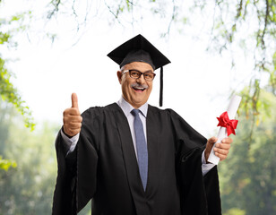 Man in a graduation gown holding a diploma and showing thumbs up outdoors with trees behind