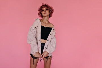 Wall Mural - Curly pink-haired woman in dark denim shorts and stylish oversized jacket poses on pink background. Attractive tattooed girl in sunglasses looks gorgeous .