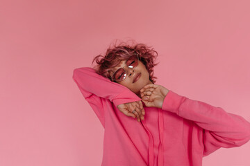 Wall Mural - Portrait of tanned curly woman in sunglasses and pink hoodie smiles on isolated. Cool attractive girl poses on pink background.