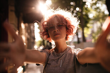 Poster - Curly short-haired woman in grey tee takes selfie. Joyful girl in sunglasses looks into camera outside on street.