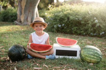 Wall Mural - Kid with hat smiling very emotional and eating watermelon. Funny child hold fruit. Outdoors in park, summers season of fruits and berries. healthy food