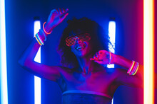 Fashionable Woman With Afro Hairstyle Dancing In Room With Colorful Lamps. Cyberpunk Style Or Hipster With Fluorescent Bracelets And Glasses. Night Club, Futuristic Outfit. Sexy Lady Concept. 