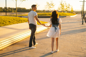  Back view of young couple in love holding hands walking in city park in sunny summer day on background of bright sunlight. Happy relationships between boyfriend and girlfriend enjoying time together.