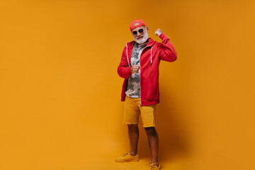 Adult man in street style outfit demonstrates muscles. Stylish bearded guy with tattoos in red hoodie and orange shorts posing..