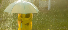 A Happy Girl With A Green Umbrella Under The Summer Rain. The Girl Is Dressed In A Yellow Raincoat And Enjoys The Rain. Banner. The Child Plays In Nature In The Fresh Air.