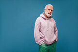 Charming man in white hoodie looking into camera on blue background. Serious bearded guy with grey hair in lilac clothes posing..