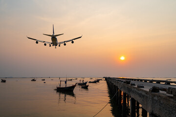 rear image commercial passenger aircraft or cargo airplane fly over fishing wood boat floating in the sea at jetty in evening with golden sunset seascape view