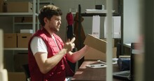 Man In Red Vest, Employee Of Warehouse Taking Photo Of Parcel On Smartphone, Writing Information About Delivery Box On The Background Of Cardboard Boxes. Logistics, Delivering And Storage Concept. 