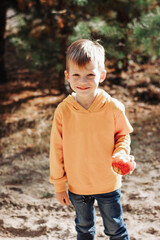 Portrait of little handsome smiling boy in orange hoodie and jeans holds red apple in the hand while walking in pine forest at sunny day. Vertical orientation 