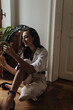 © Look! - Happy black haired lady in white shirt, wide pants and dark shoes looking at cute flower and sitting on floor in apartment..