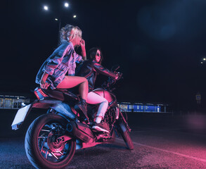 a couple of girls in love on a motorbike in the rays of neon light in an empty parking lot at night