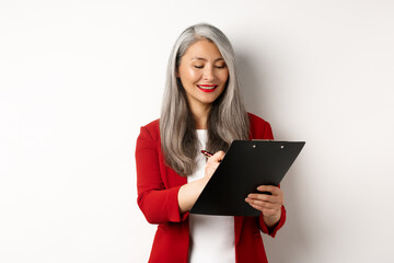 Successful asian senior businesswoman with grey hair taking notes on clipboard, inspecting enterprise, standing over white background