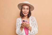 Pleased Dreamy Young Asian Woman Holds Modern Smartphone In Hands Uses Cellular Application For Chatting Online Looks Away Wears Stylish Hat White Knitted Shawl Isolated Over Brown Background