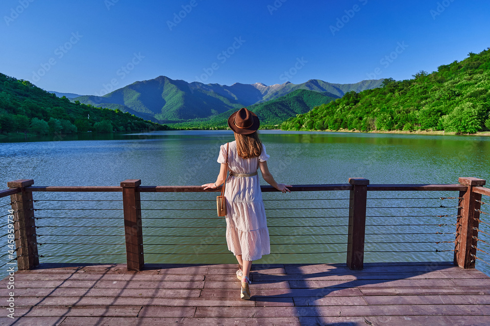 Traveler girl standing alone on edge of pier and staring at lake and mountains. Enjoying beautiful freedom moment life and serene quiet peaceful atmosphere in nature. Back view - obrazy, fototapety, plakaty 