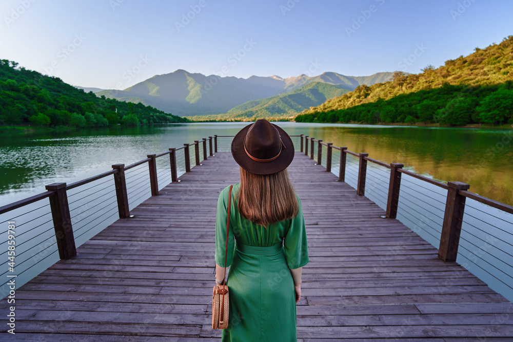 Traveler girl standing alone on pier and staring at lake and mountains. Beautiful freedom moment and serene peaceful atmosphere in nature. Back view - obrazy, fototapety, plakaty 