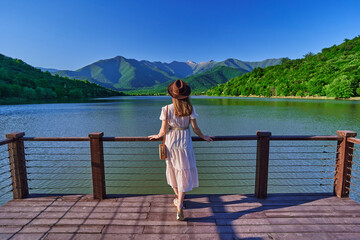 Wall Mural - Traveler girl standing alone on edge of pier and staring at lake and mountains. Enjoying beautiful freedom moment life and serene quiet peaceful atmosphere in nature. Back view