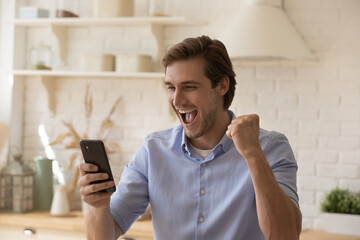 Wall Mural - Overjoyed young male sit at kitchen scream yes raise clenched fist up looking on phone screen celebrate getting great news. Excited joyful millennial guy make winner gesture read message about reward