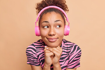 Wall Mural - Close up portrait of thoughtful pleased dark skinned woman keeps hands under chin wears headphones on ears listens audio track or tutorial lecture concentrated aside isolated over beige wall