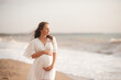 © morrowlight - Smiling beautiful pregnant woman wear stylish white dress hold belly walk at beach over waves of sea outdoors. Looking away. Motherhood. Maternity. Healthy lifestyle.
