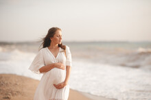 Smiling Beautiful Pregnant Woman Wear Stylish White Dress Hold Belly Walk At Beach Over Waves Of Sea Outdoors. Looking Away. Motherhood. Maternity. Healthy Lifestyle.