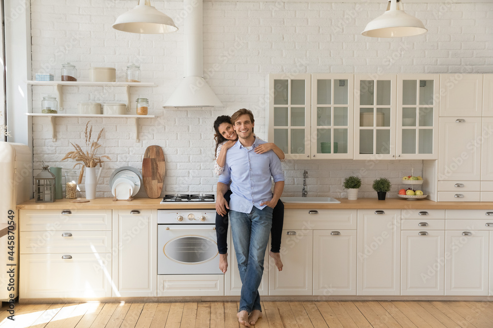 Portrait of affectionate young wife sitting on kitchen counter hug smiling husband from behind at modern renovated interior. Loving young couple homeowners look at camera glad of buying new furniture - obrazy, fototapety, plakaty 