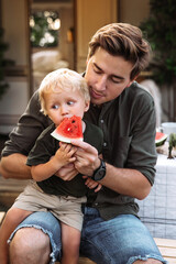 Wall Mural - Small boy eat watermelon sit on father knees outside house in garden or near camper car while road trip vacation. Young dad feed baby son with slice of fruit outdoors. Loving male parent caring of kid