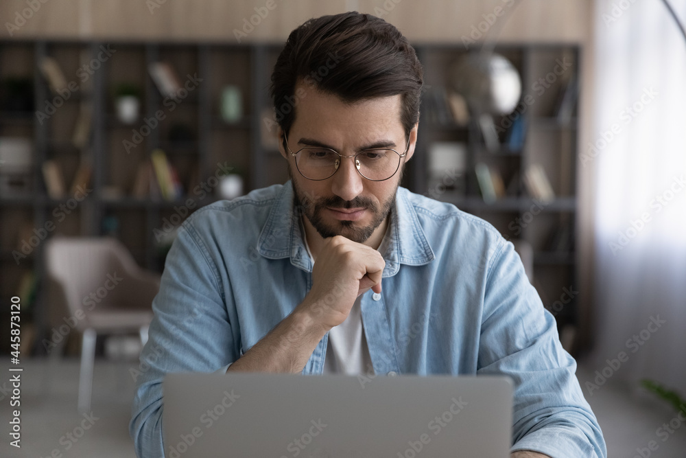 Thoughtful young male manager in eyeglasses looking at laptop screen, considering problem solution, preparing research report, analyzing sales statistic, developing strategy, working on online project - obrazy, fototapety, plakaty 