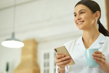 Wall Mural - Happy female doctor standing with smartphone and cup